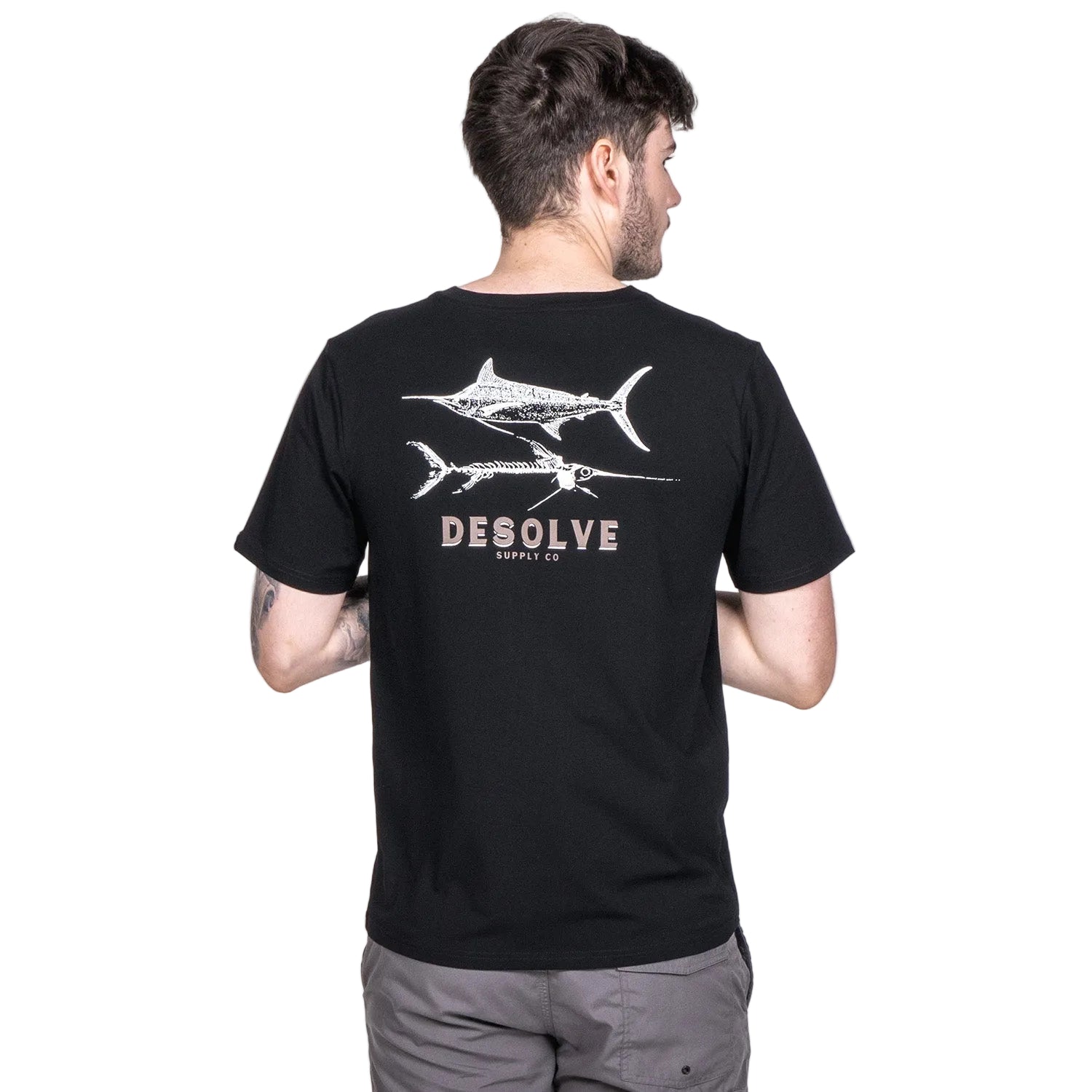 A person wearing a black crew neck t-shirt with a white graphic of two sharks on the front and the text 'DESOLVE' below it. There is a 'UP50+' label at the bottom left corner.